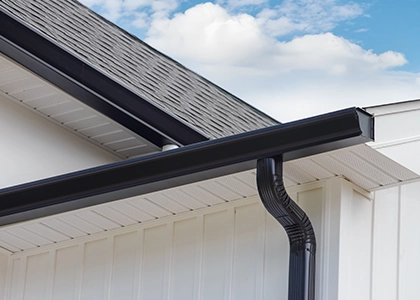 A close-up of a black metal gutter and downspout attached to the edge of a white house, with a bright blue sky and clouds visible in the background.